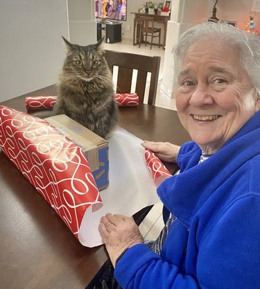 Woman laughing at cat interrupting her present wrapping by sitting on the paper.